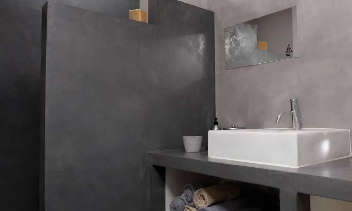 Salle de bain moderne avec murs en béton ciré. Une douche sans joint, un lavabo rectangulaire et des serviettes pliées créent une ambiance épurée.
