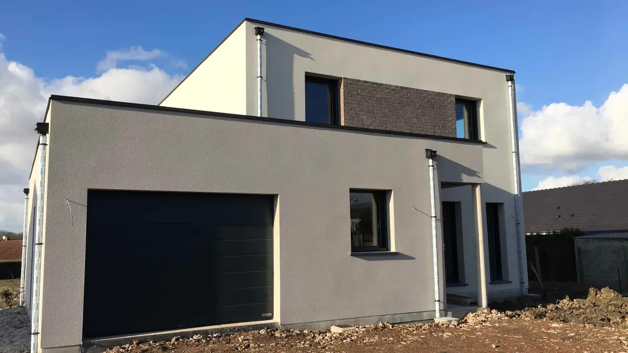 Maison moderne avec garage intégré. La couleur façade maison est un gris clair, contrastant avec des touches de blanc cassé et un bandeau de briques sombres.