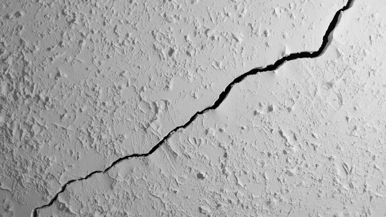 Gros plan sur une fissure traversant un plafond texturé. La fissure est sombre et profonde, contrastant avec la surface blanche et irrégulière.