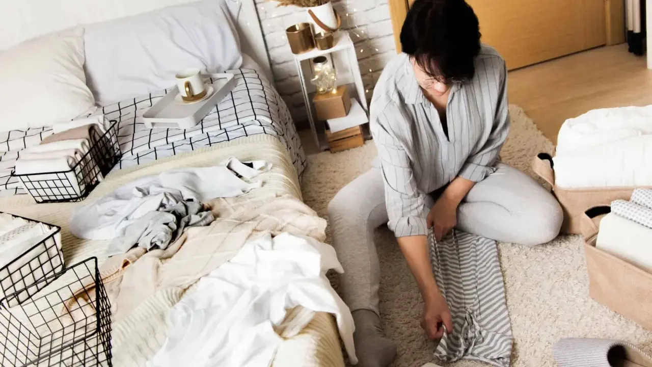 Une femme plie du linge sur un tapis, à côté d'un lit défait. Elle apprend comment ranger sa chambre adulte efficacement.