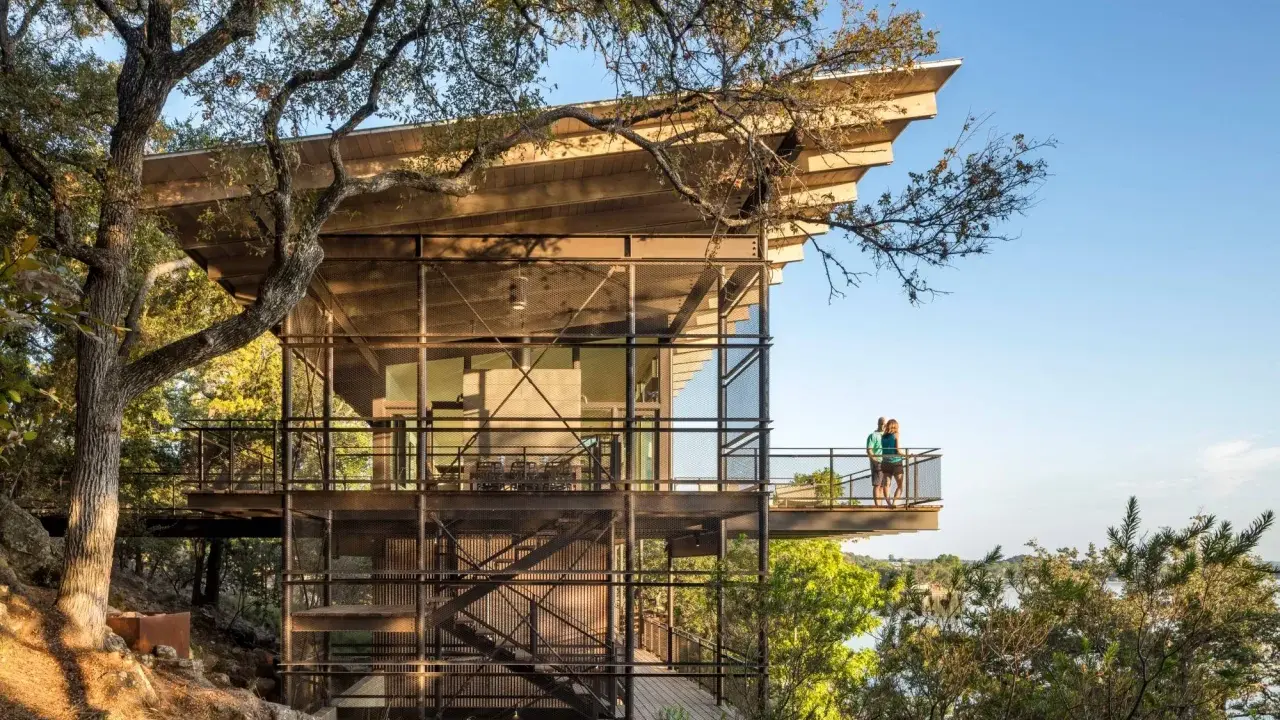 Maison moderne sur un terrain en pente raide, avec des balcons et des escaliers extérieurs en métal, nichée dans les arbres près d'un lac.