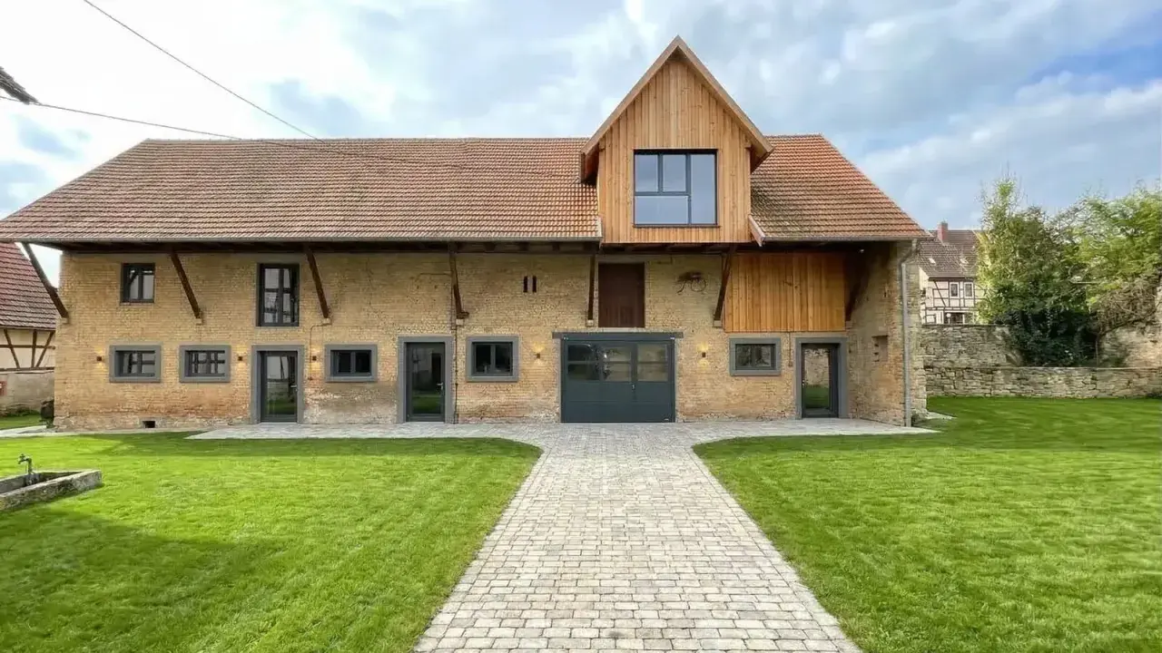 Belle grange rénovée avec une façade en briques jaunes, un toit en tuiles et un ajout moderne en bois. Un chemin pavé mène à l'entrée principale.