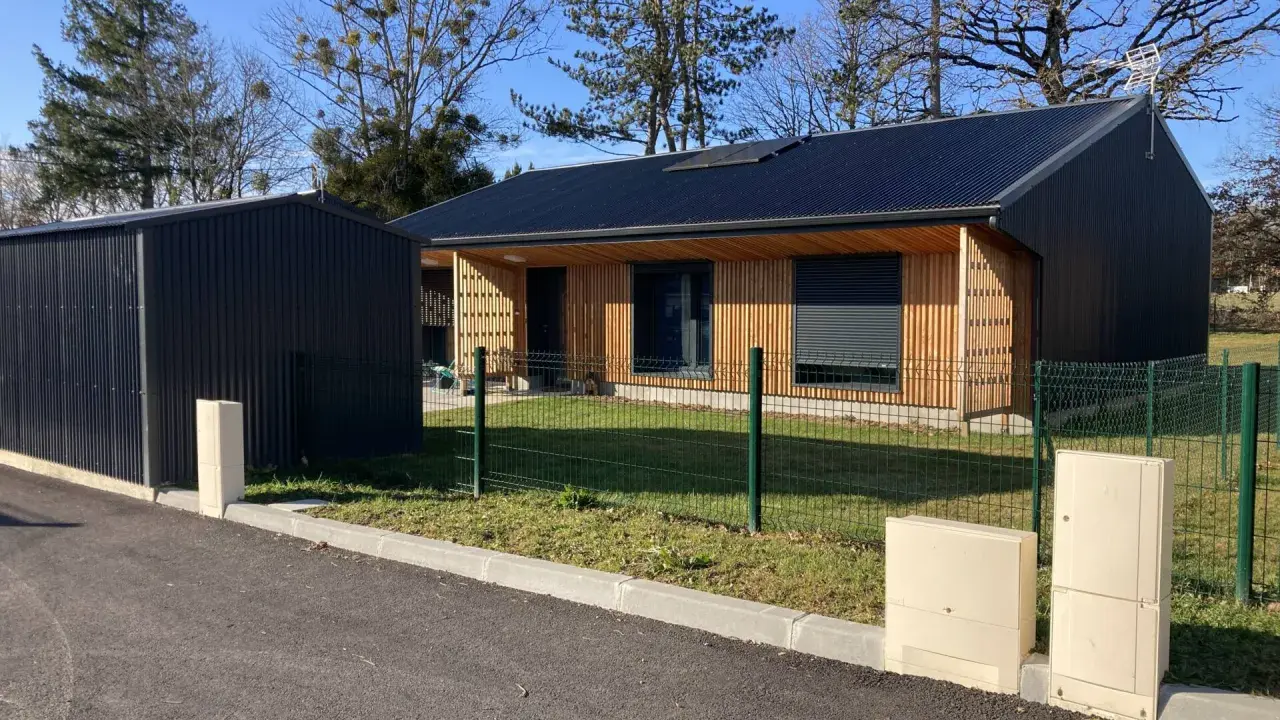 Maison passive ossature bois avec panneaux solaires sur le toit, bardage bois et métal, devant un ciel bleu et des arbres.
