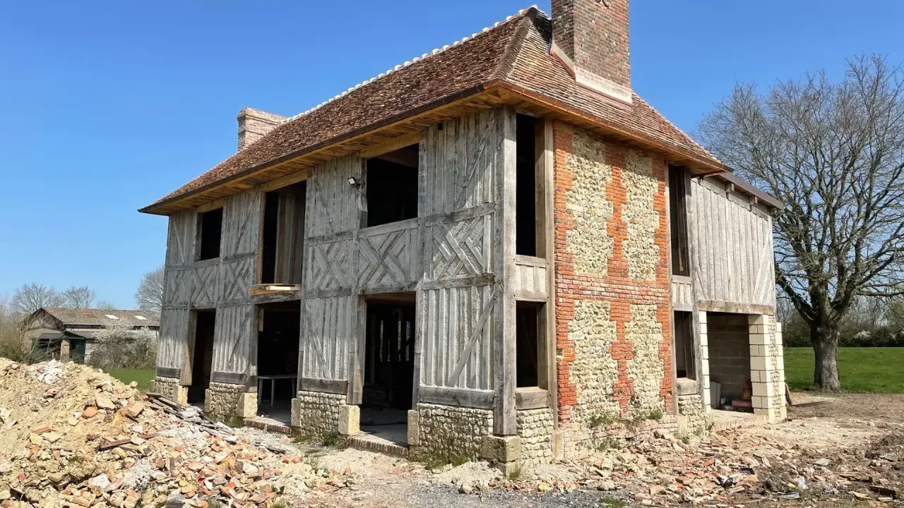 Construction d'une maison normande traditionnelle, avec colombages apparents et murs en pierre et brique. Le chantier est en cours.