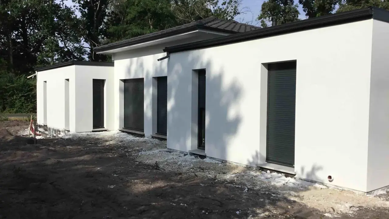 Maison moderne avec toit plat, plan de 150m2. Construction blanche aux lignes épurées, entourée d'arbres.