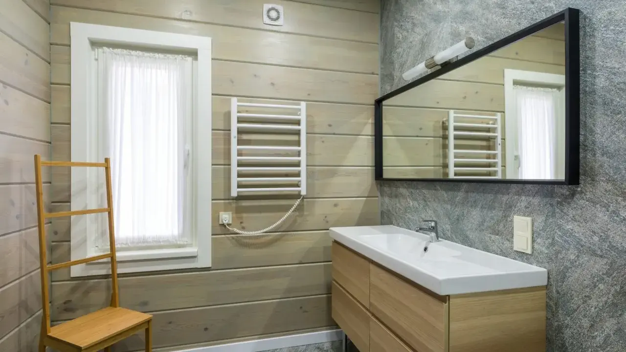 Salle de bain moderne avec murs en bois clair et pierre grise. Un meuble vasque en bois, un miroir, un radiateur et une échelle en bambou pour habiller un mur.