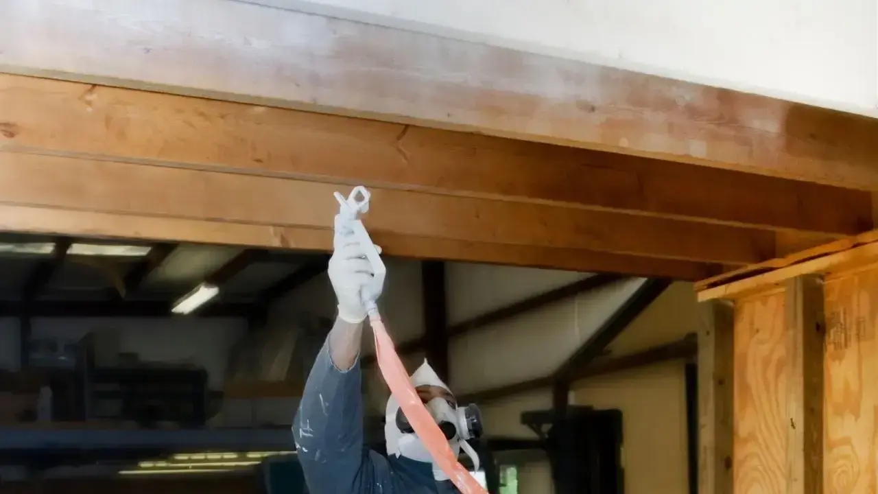 Un ouvrier masqué et ganté applique une peinture blanche sur une poutre en bois, transformant l'aspect rustique.