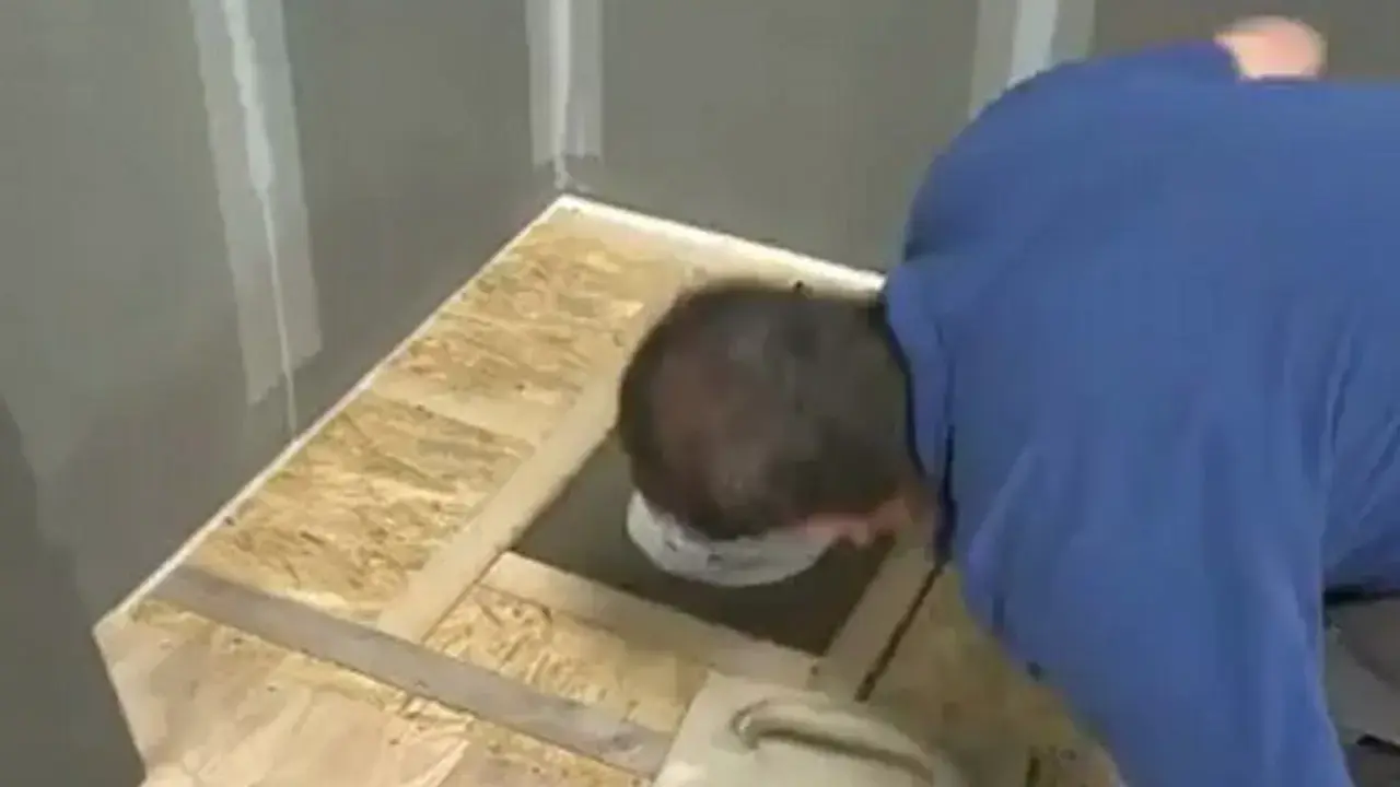 Un homme installe une douche sur un plancher en bois brut. Il utilise une truelle pour étaler du mortier autour du drain.