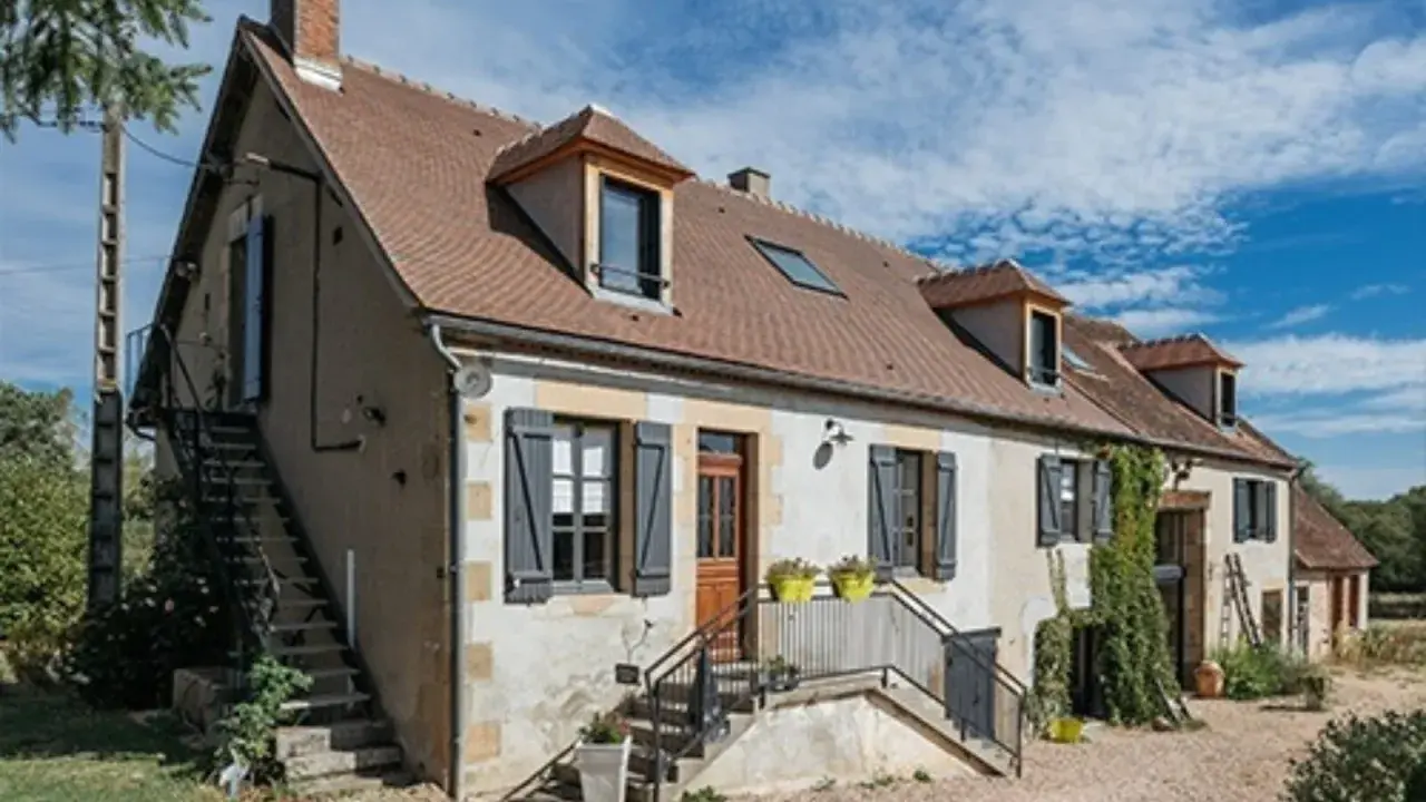 Une maison de campagne simple avec des volets gris, une cheminée et des escaliers extérieurs sous un ciel bleu.