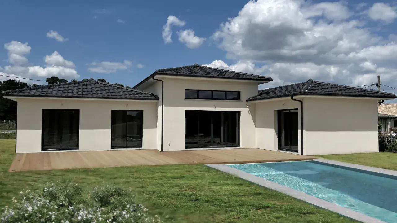 Une maison du sud moderne avec piscine, terrasse en bois et grand jardin sous un ciel bleu nuageux.
