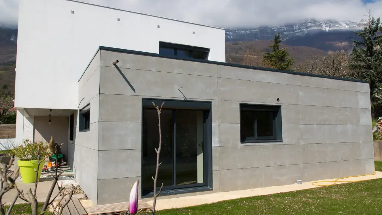 Extension moderne en parpaing gris, 20m2, avec grandes baies vitrées et vue sur montagnes.