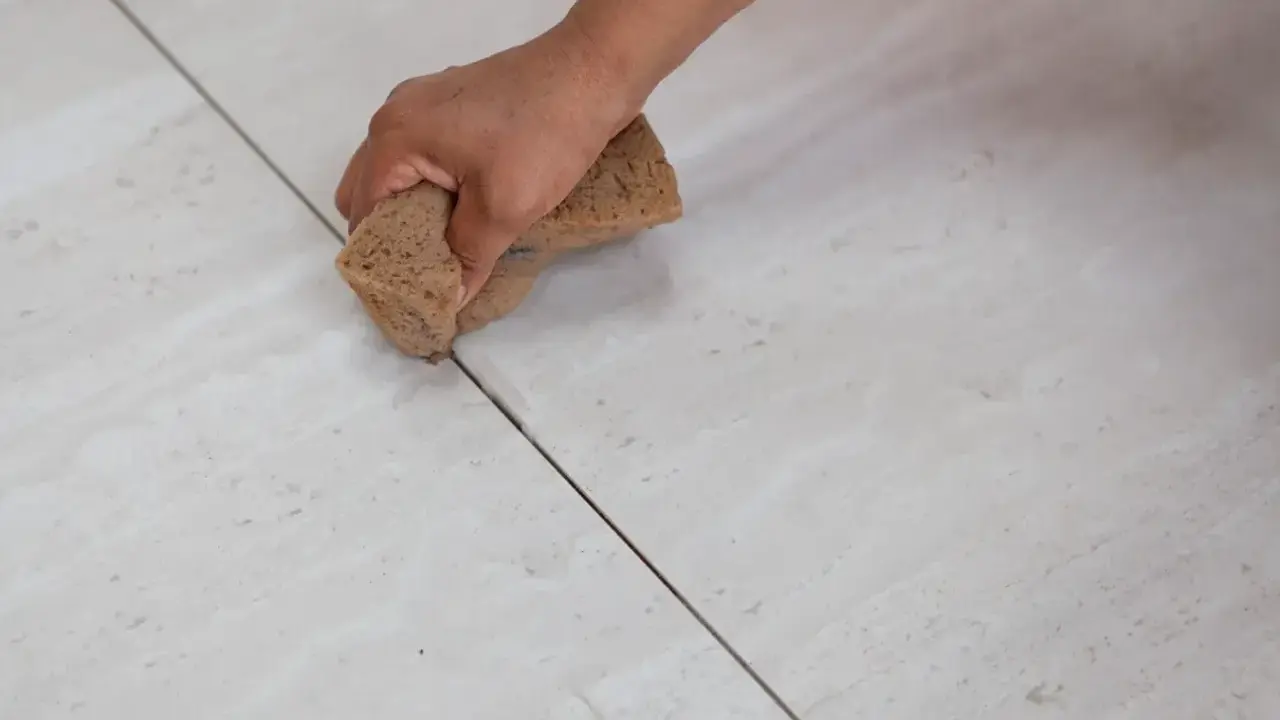 Main d'une personne nettoyant le joint de carrelage avec une éponge. La couleur du joint est sombre, contrastant avec le carrelage clair.