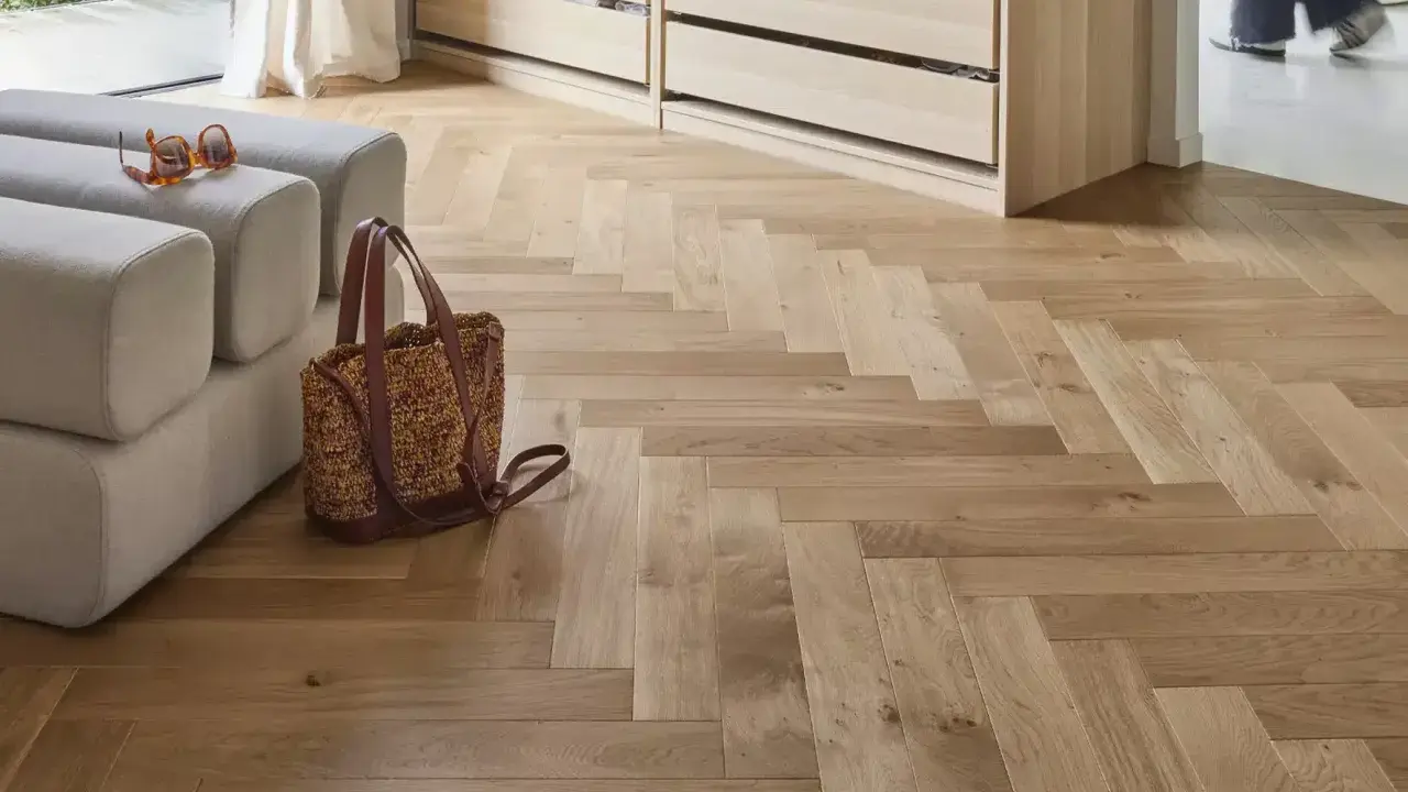 Un parquet en bois clair au calepinage en chevrons, avec un canapé, un sac et des lunettes posés dessus.