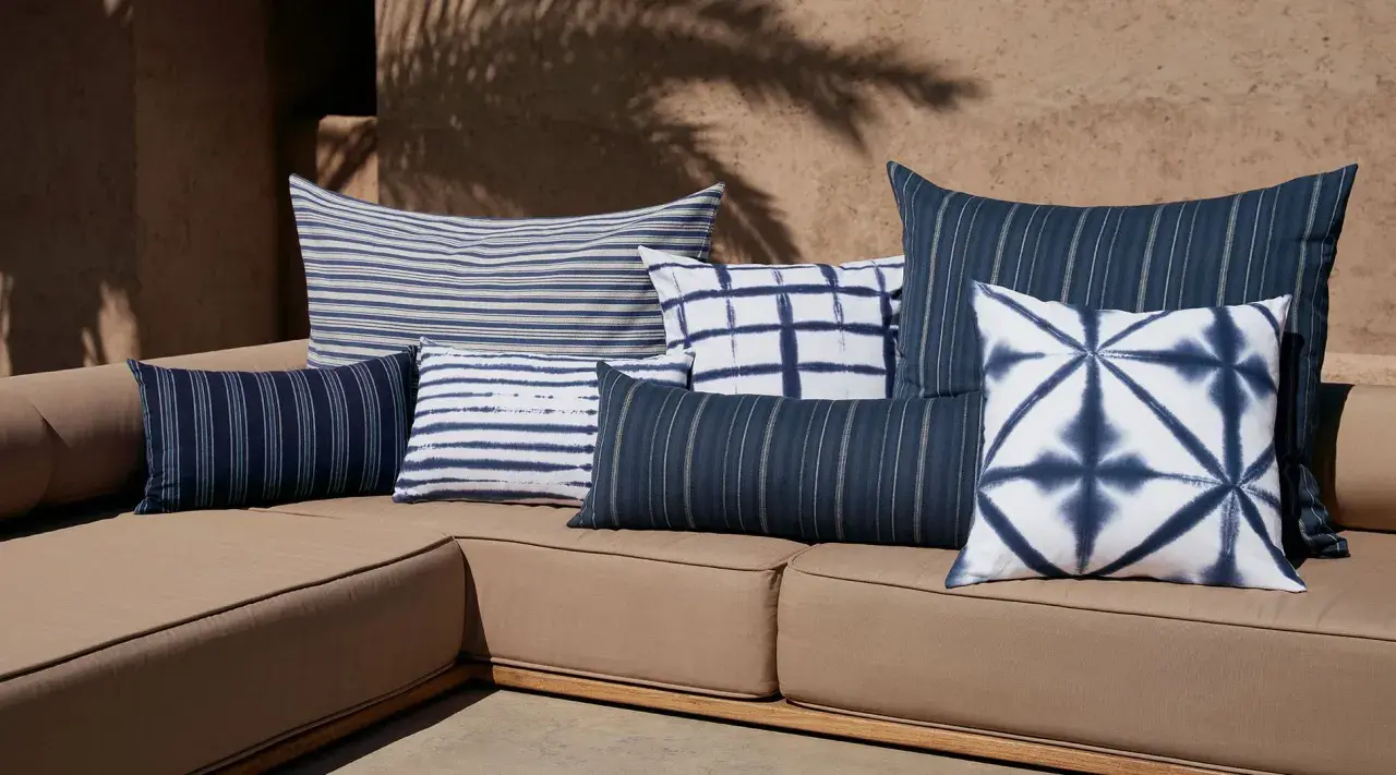 Plusieurs coussins bleus et blancs, à rayures et motifs tie-dye, sont disposés sur un canapé marron. Idée pour comment mettre un plaid sur un canapé.