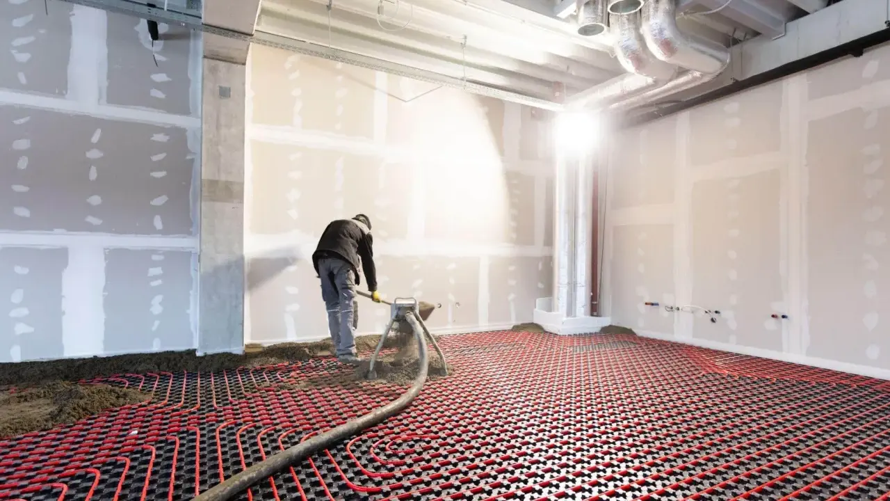 Un ouvrier installe un système de chauffage au sol hydraulique. Des tuyaux rouges serpentent sur une grille noire, prêts à diffuser une chaleur douce.