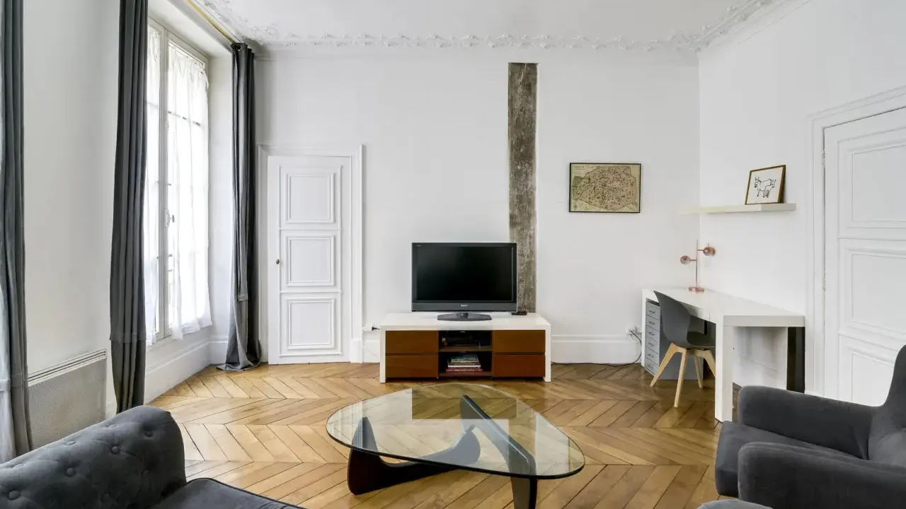 Salon moderne avec canapé gris, table basse en verre, bureau et mur blanc orné d'une carte de Paris.