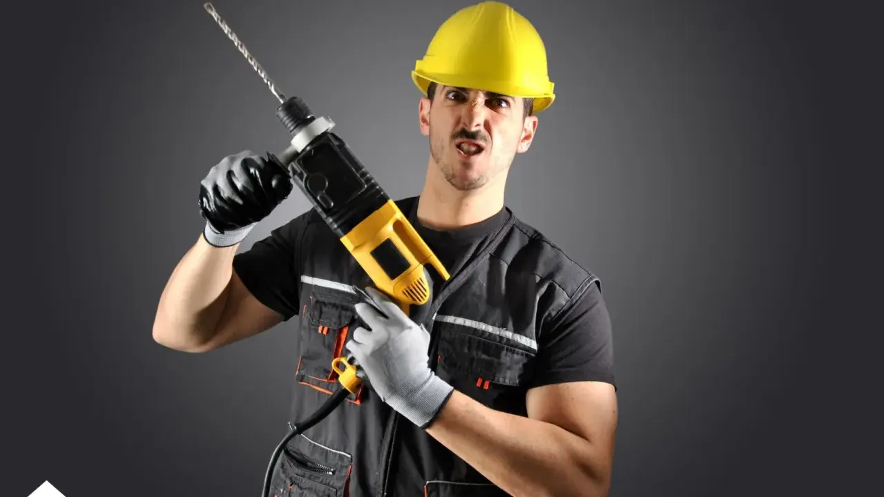 Un homme en casque jaune, gants et tenue de travail tient une perceuse. Il a une expression faciale exagérée, comme un architecte ou un constructeur prêt à relever un défi.