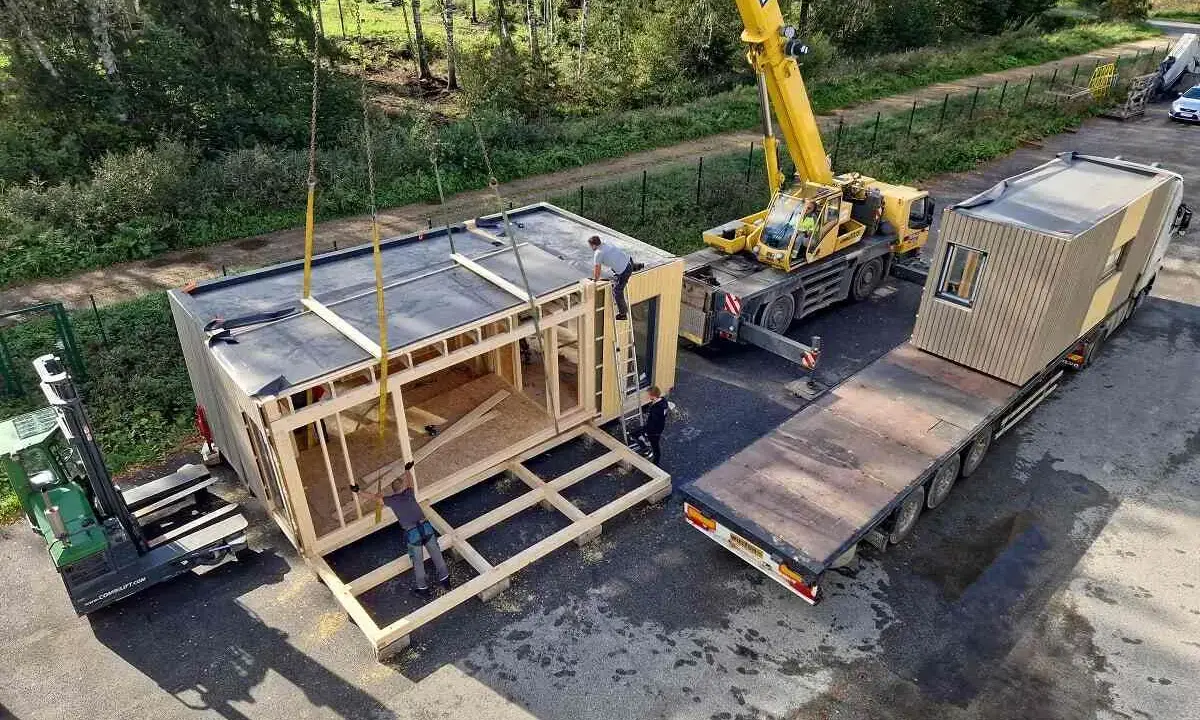 Une grue soulève un module de construction pour des maisons modulaires. Un chariot élévateur et un camion plateau sont également présents.
