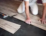 Pose de parquet flottant : un artisan installe des lames de bois sur un sol noir. Le prix de pose est une préoccupation pour ce type de revêtement.
