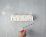 Main tenant, une main tient un rouleau à peinture blanc sur un mur bleu clair. Les traces de rouleau à la lumière révèlent la texture du mur.