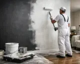 Un peintre en salopette blanche est en train de repeindre un mur de couleur foncée en blanc, à l'aide d'un rouleau.
