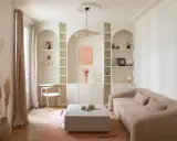 Un coin bureau discret dans un salon cosy, avec une étagère murale arche, un canapé beige et un tapis rose.