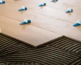 Pose de carrelage 60x60 en cours. Des carreaux en bois clair sont posés sur du mortier frais, avec des croisillons bleus et blancs pour un espacement parfait.