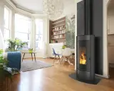 Un poêle à bois moderne avec un parement texturé derrière, dans un salon lumineux avec des plantes et une bibliothèque.