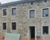 Maison ancienne en cours de rénovation, avec des murs en pierre brute et de nouvelles fenêtres. Le projet de construire en pierre continue.