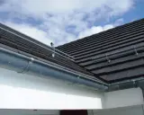 Le chêneau de cette maison, en zinc, est bien fixé sous le toit de tuiles sombres. Le ciel bleu et nuageux promet une belle journée.