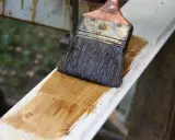 Application d'une lasure bois pour un avant-après visible. Le pinceau dépose une teinte chaude sur le bois brut.