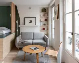 Aménagement petit appartement : lit surélevé, canapé gris, table basse ronde en bois, étagères et grande fenêtre lumineuse.