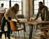 Trois personnes arrangent des chaises autour d'une grande table en bois brut. Quelles chaises avec table industrielle, le tout dans un loft lumineux.