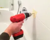 Une main tient une perceuse rouge pour percer du carrelage blanc, avec du ruban adhésif pour guider le foret.