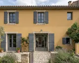 Façade d'une maison provençale à l'enduit projeté ocre, avec volets gris et végétation luxuriante.