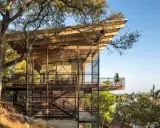 Maison moderne sur un terrain en pente raide, avec des balcons et des escaliers extérieurs en métal, nichée dans les arbres près d'un lac.