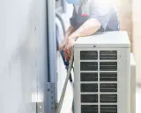 Un technicien s'occupe de recharger la clim de la maison. Il manipule des tuyaux près de l'unité extérieure.