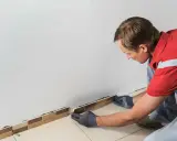 Un homme en tenue de travail enfile des gants pour enlever la plinthe du carrelage.