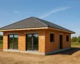Une petite maison à construire en briques orange, avec un toit sombre et de grandes fenêtres, sur un terrain sablonneux sous un ciel bleu.