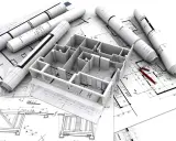 Maquette 3D d'une maison familiale sur des plans d'architecte, avec des rouleaux de plans et des stylos rouges.
