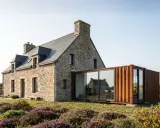Maison en pierre avec une extension moderne en container maritime vitrée, offrant une vue sur la campagne fleurie.
