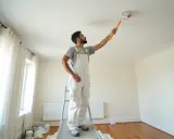 Peintre en bâtiment sur un escabeau, appliquant de la peinture au rouleau sur un plafond. Il est difficile de dire combien de temps pour peindre un plafond de 50m2.