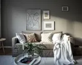 Salon cosy avec un **canapé gris clair** orné de coussins texturés et d'une couverture blanche. Des œuvres d'art abstraites décorent le mur.