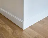 Un coin de mur blanc avec des plinthes moulurées et un parquet en bois clair. Ces plinthes servent à protéger les murs et à cacher les imperfections.