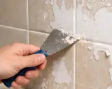 Main tenant enlever peinture sur carrelage avec une spatule. La peinture s'écaille sur les carreaux beiges.
