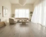 Un salon lumineux avec un canapé incurvé beige, une table basse en pierre et un sol en bois gris clair. Le beige et le blanc s'accordent parfaitement avec le gris clair.