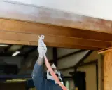 Un ouvrier masqué et ganté applique une peinture blanche sur une poutre en bois, transformant l'aspect rustique.