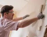 Un homme avec des lunettes de protection et des gants commence à décoller du vieux papier peint d'un mur, une femme l'observe.