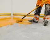 Un ouvrier en combinaison orange et grise, les bottes sales, utilise un rouleau pour peindre le sol d'un garage en jaune vif.