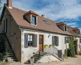Une maison de campagne simple avec des volets gris, une cheminée et des escaliers extérieurs sous un ciel bleu.